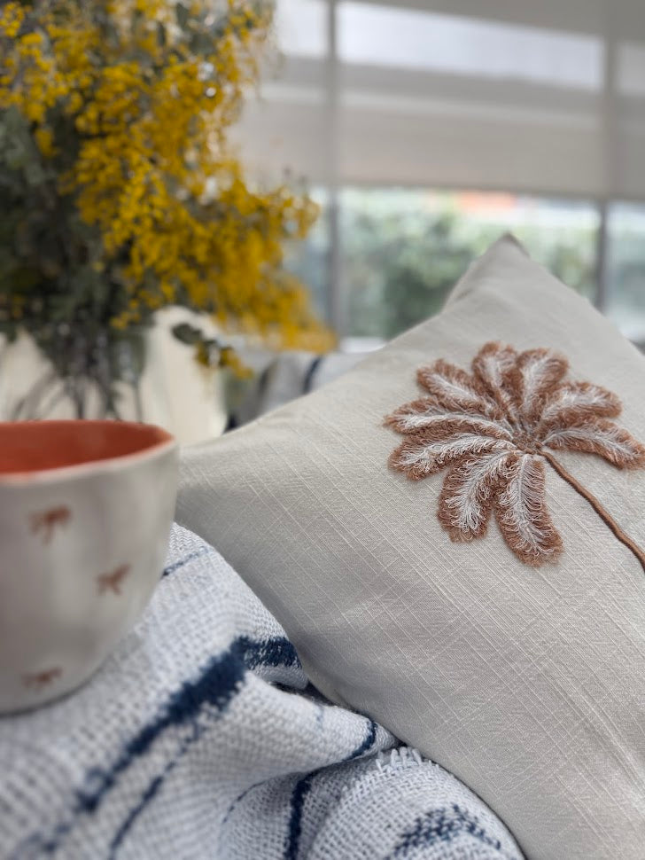 Cushion with palm tree embroidery on a couch next to a cup and blanket