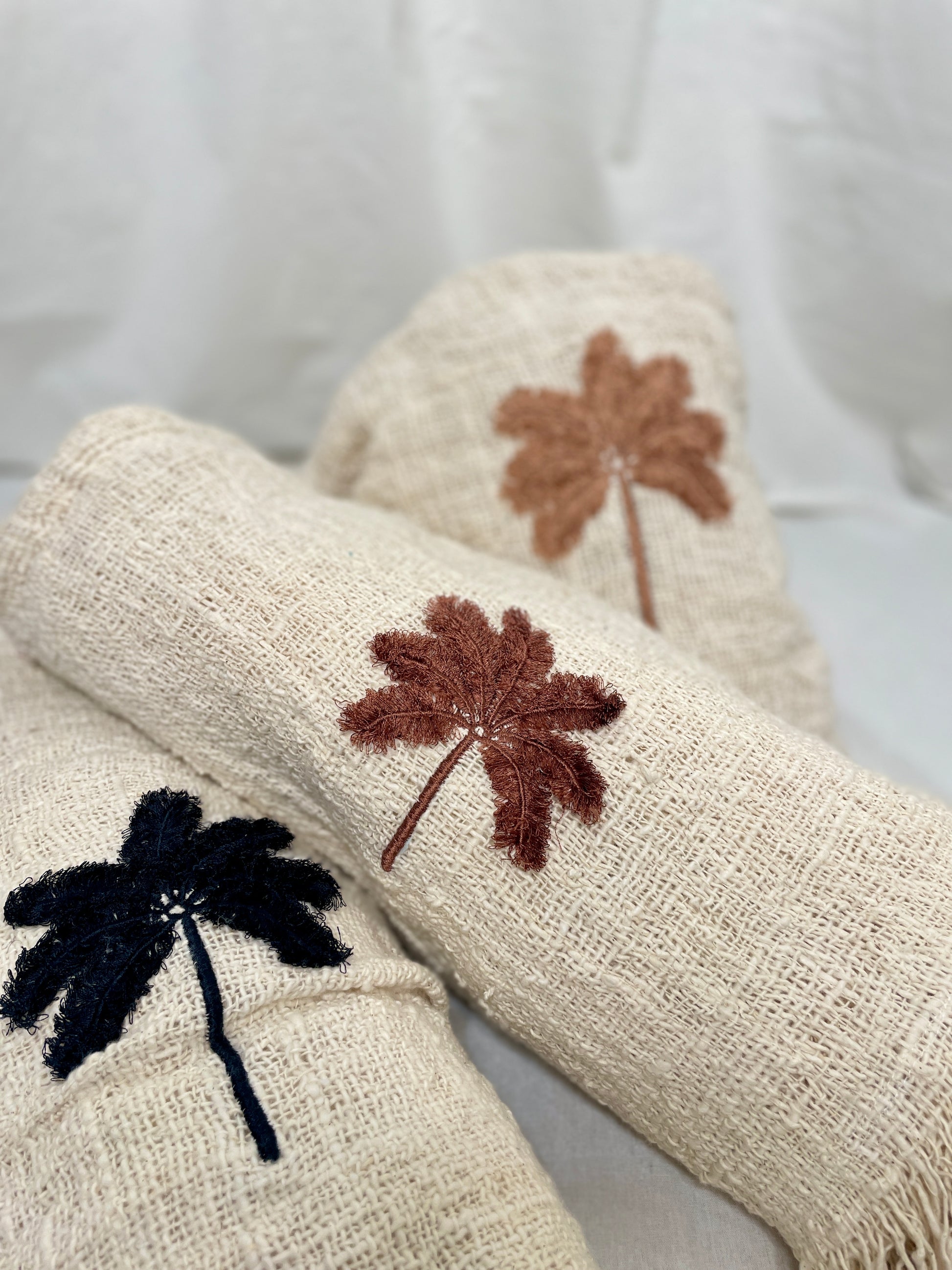 Three natural linen throws rolled up, each with a hand embroidered palm tree in different coloured