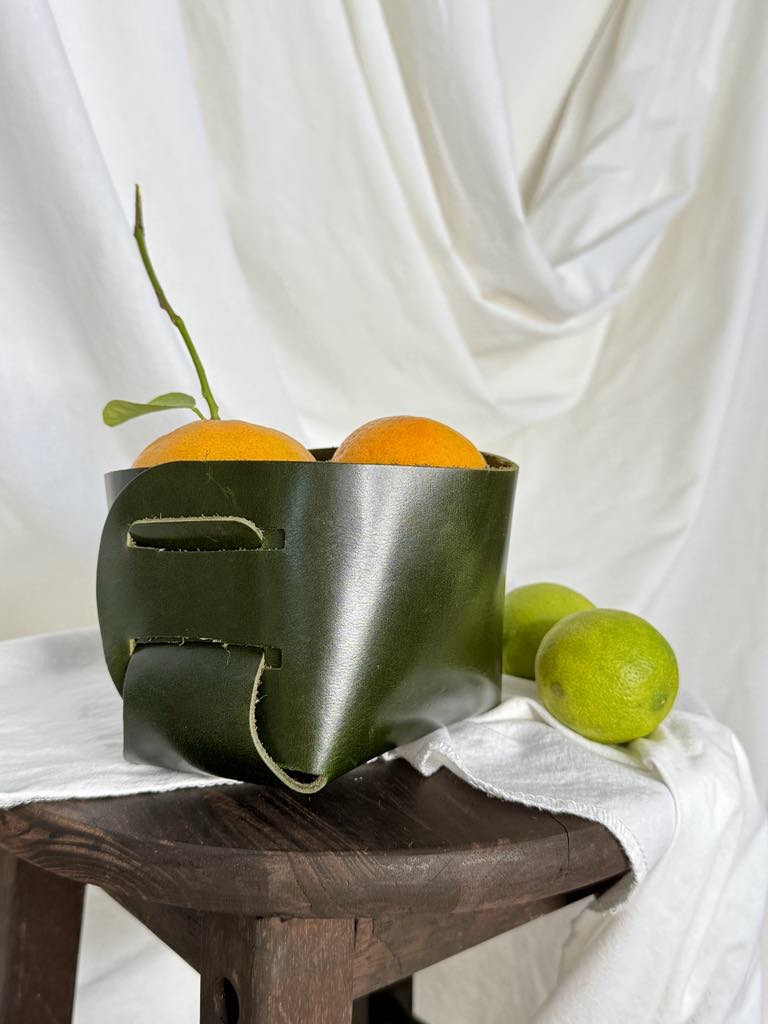 Green leather fruit basket with oranges and limes on a white fabric background