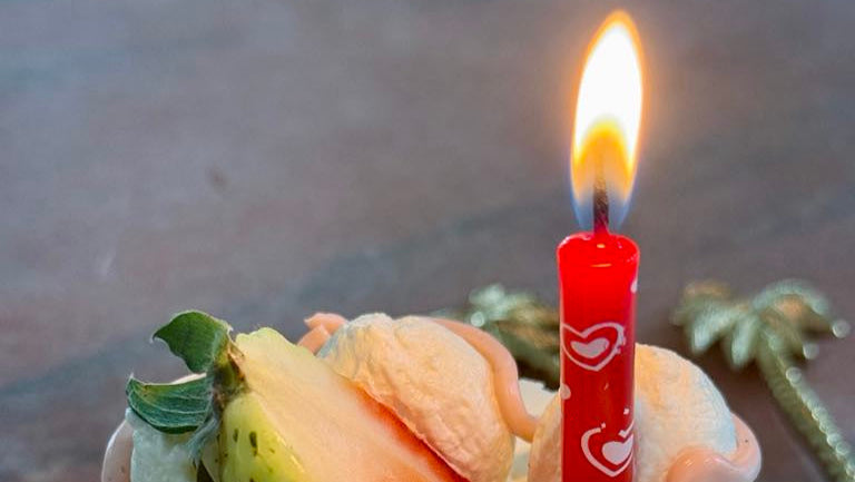 Small birthday cake with a lit candle, fruit slices, and white frosting on a reflective surface.