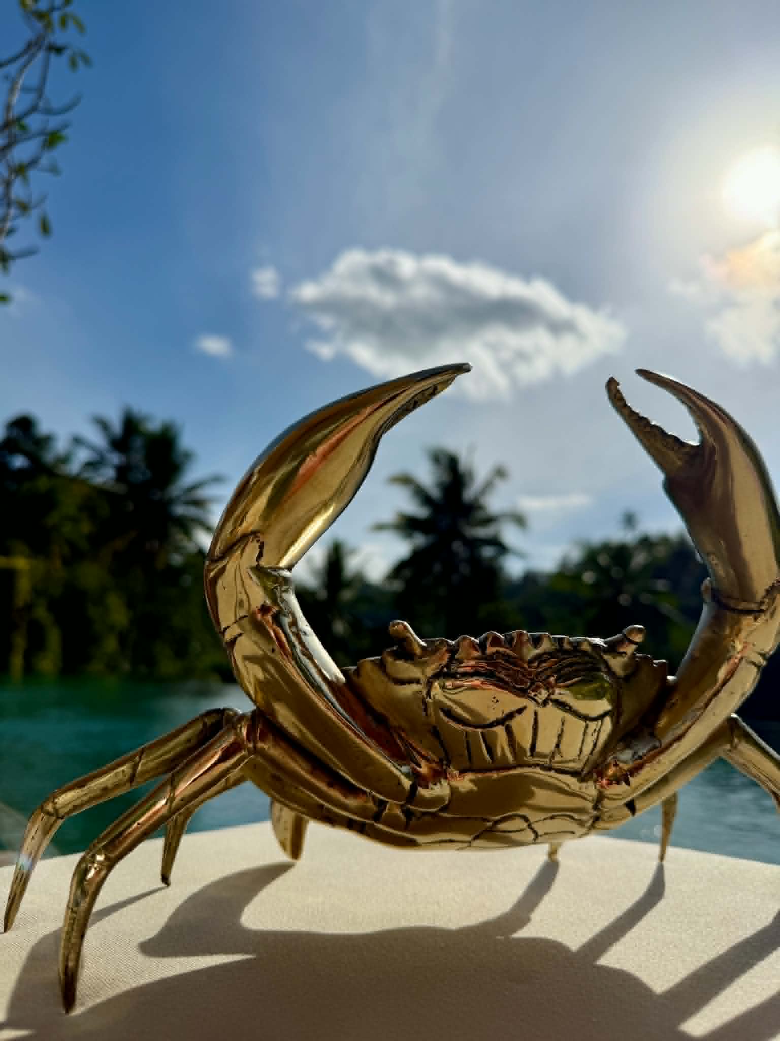 A shiny metallic gold crab figurine or sculpture displayed outdoors on a light surface, with a blurred tropical background featuring palm trees, blue water, and bright sunlight.