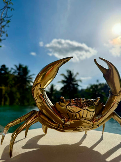 A shiny metallic gold crab figurine or sculpture displayed outdoors on a light surface, with a blurred tropical background featuring palm trees, blue water, and bright sunlight.