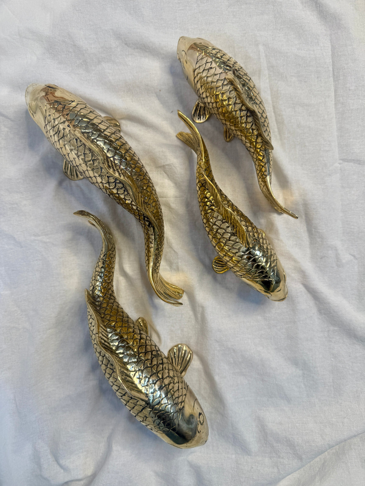 Four brass koi fish ornaments with detailed scales, displayed on a wrinkled white fabric background.