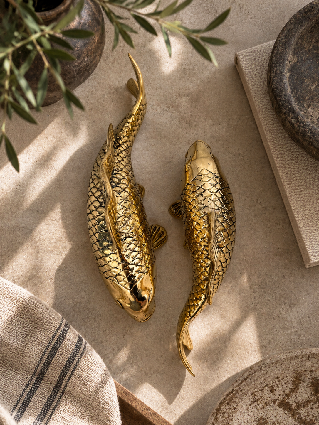Two brass fish-shaped wall decor sculptures with detailed scale patterns, displayed on a neutral beige stone surface with rustic props.