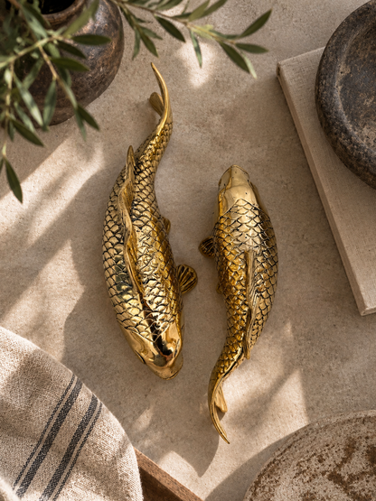 Two brass fish-shaped wall decor sculptures with detailed scale patterns, displayed on a neutral beige stone surface with rustic props.
