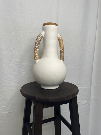 Tall pottery vase in white with two large handles either side, wrapped in rattan. Placed on a wooden stool in a studio