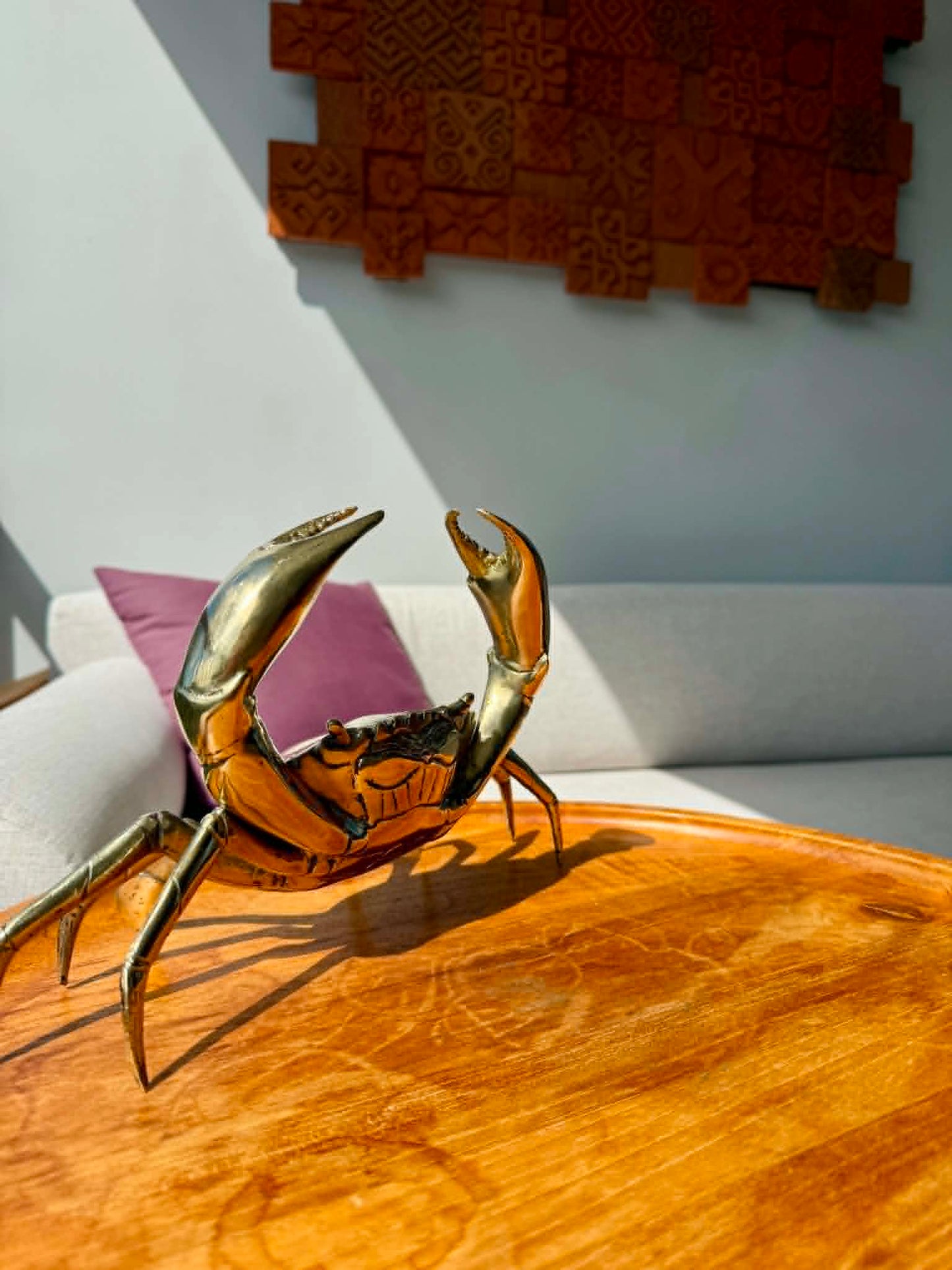 A polished brass crab figurine sculpture displayed on a round wooden table in a modern living room setting with a white sofa and textured wall art in the background.