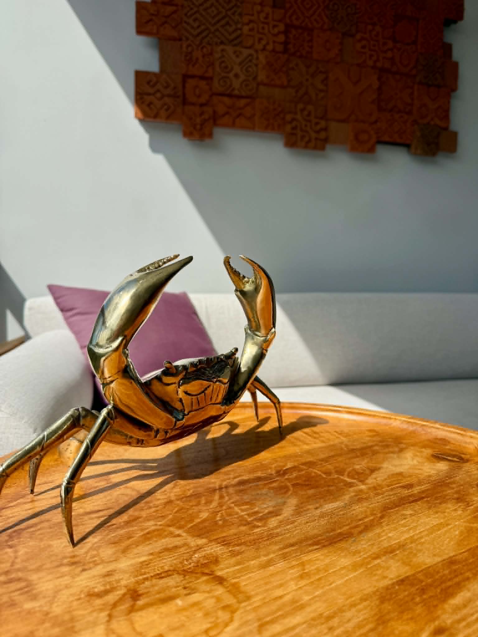A polished brass crab figurine sculpture displayed on a round wooden table in a modern living room setting with a white sofa and textured wall art in the background.