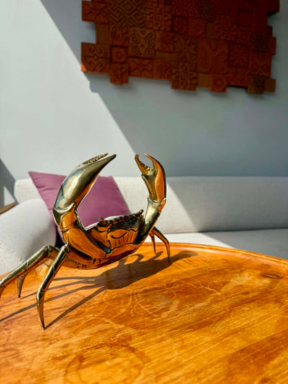 A polished brass crab figurine sculpture displayed on a round wooden table in a modern living room setting with a white sofa and textured wall art in the background.